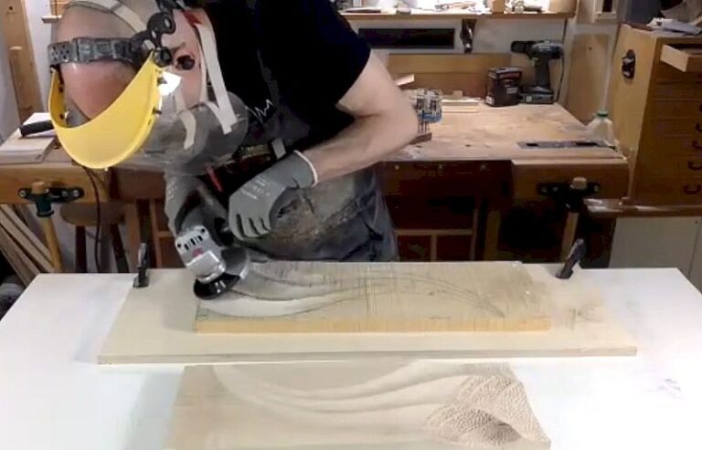 Woodworker wearing a face shield and gloves grinding a wooden block with an angle grinder on a workshop workbench, producing dust.